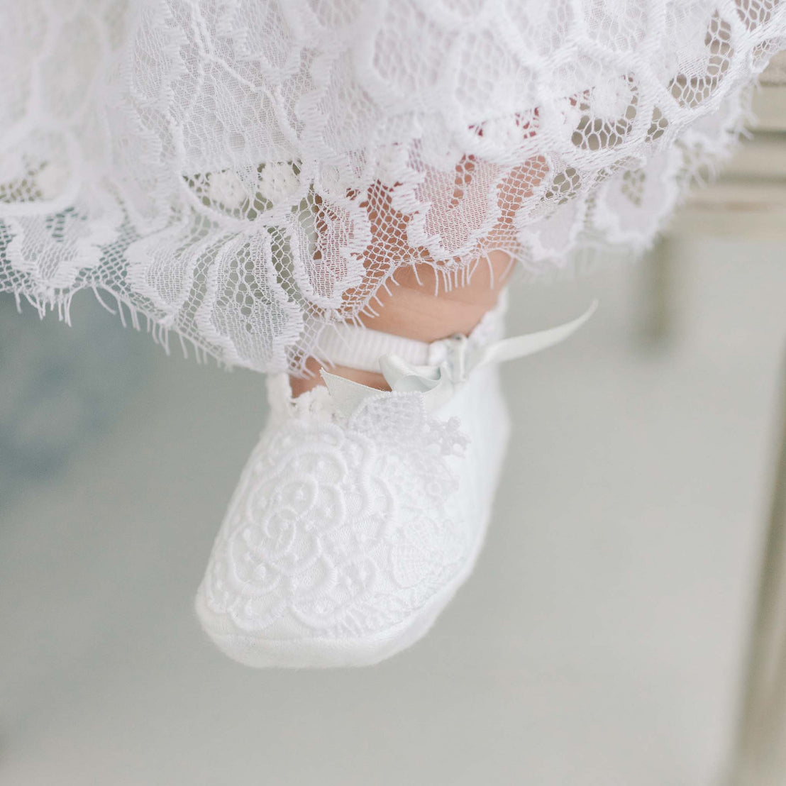 A close-up of a baby's foot adorned with a white, lace-trimmed sock and a matching Olivia Booties for a christening.