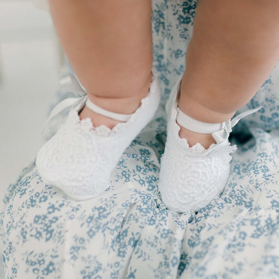 Close-up of a baby's feet in delicate Olivia Booties over a floral blue and white dress, perfect for a baptism or as a baby gift. The image emphasizes tiny details and soft textures