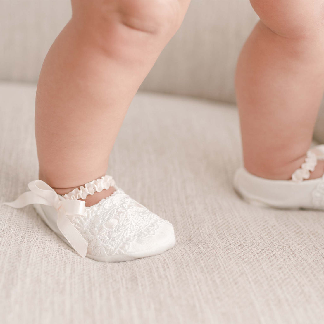Close-up of a newborn's feet in white, lace-trimmed Emma baby shoes with ribbon details, standing on a soft, beige surface.