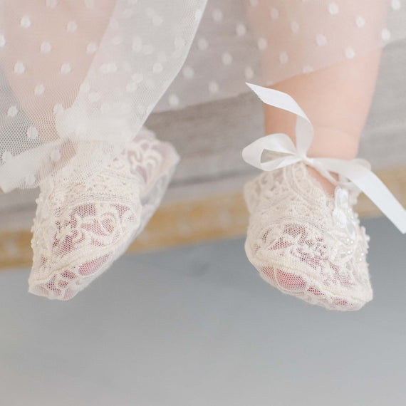 Close-up of a baby's feet in Jessica Lace Booties with white ribbon bows, suspended in the air against a softly blurred background.