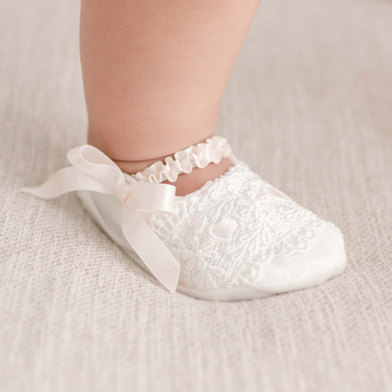 Baby foot standing on grey couch wearing white lace christening booties. 