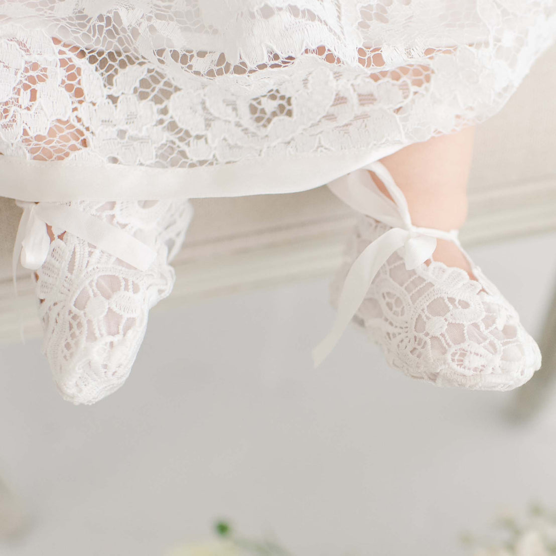 A close-up of a baby's feet in delicate white lace shoes from the Rose Accessory Bundle, tied with ribbon, barely touching the floor, with a traditional heirloom dress for a baptism.
