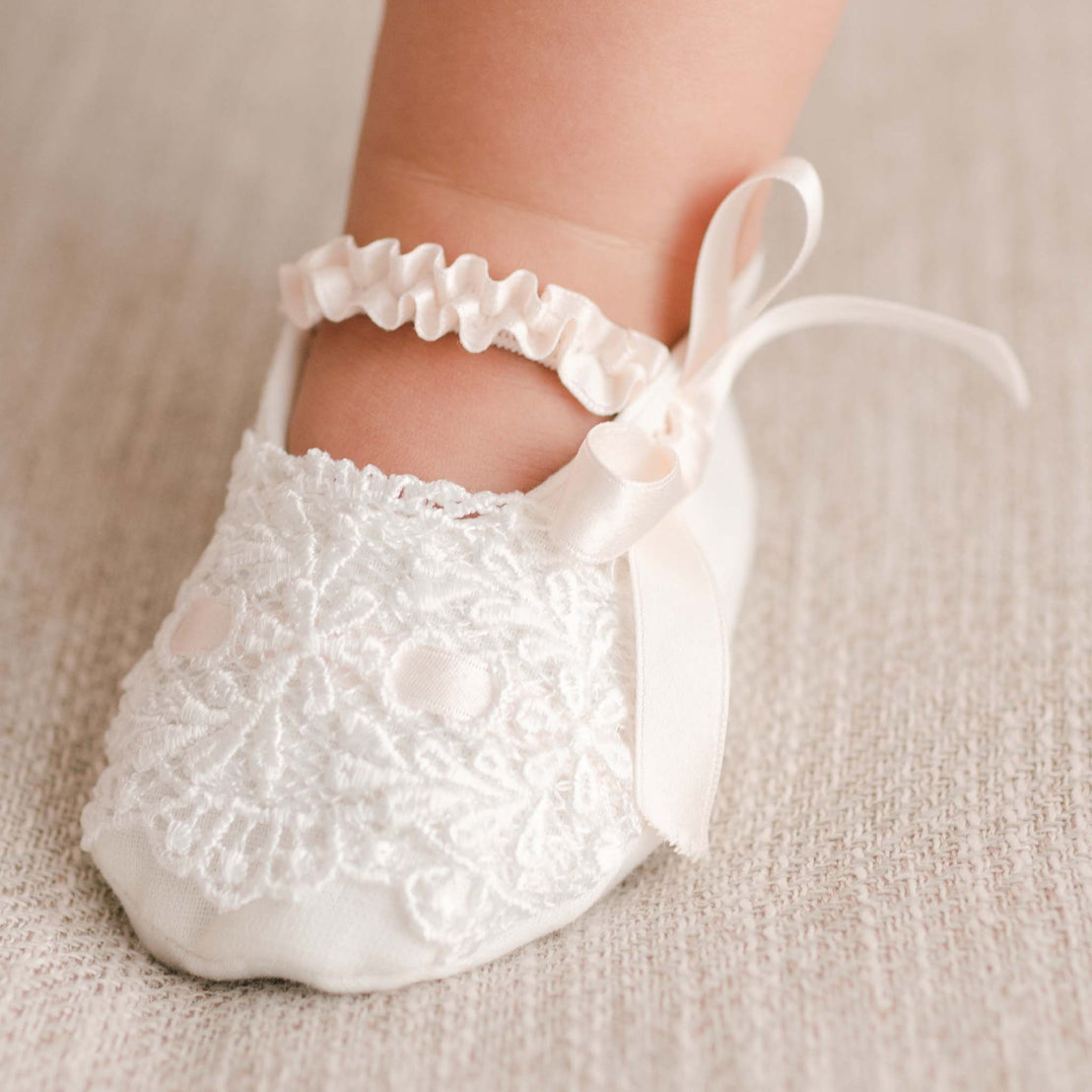A close-up image of a baby's foot adorned with a pair of Emma Booties and a frilly ankle bracelet on a soft beige background, perfect for baptism events.