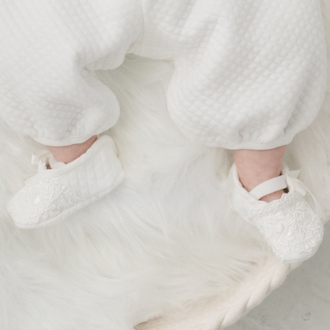 A close-up of a baby's feet wearing Madeline Quilted Booties and dressed in an upscale, textured onesie, resting on a soft white fur surface.