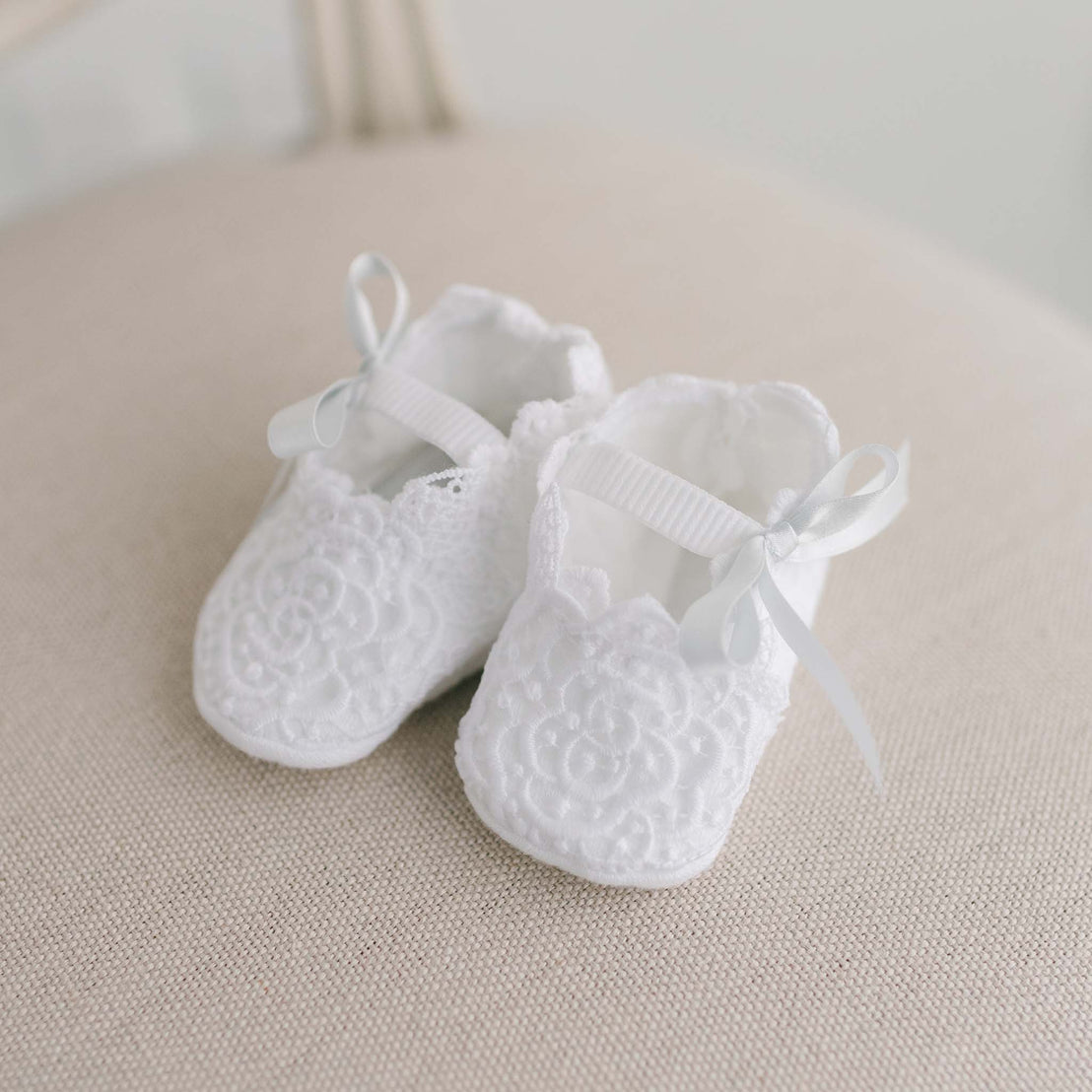 A pair of tiny, white Olivia Booties with lace details and satin ribbons, placed neatly on a soft beige surface for a christening.