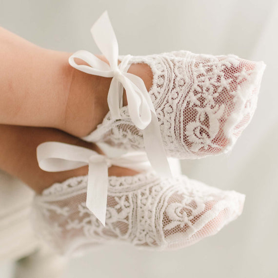 Light ivory sheer lace baby girl booties with light ivory silk ribbon ties on the feet of a baby.
