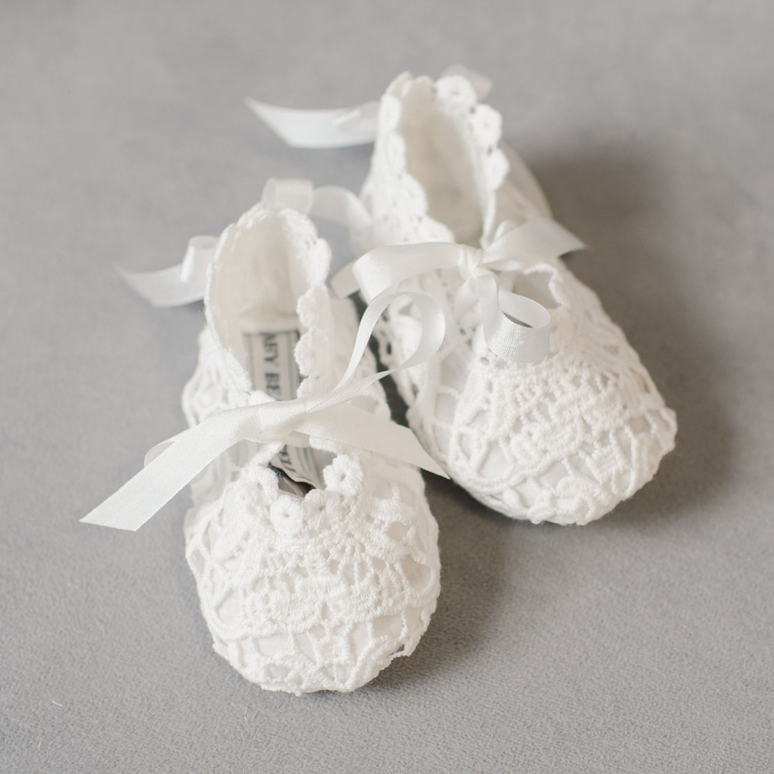 A pair of delicate Grace Lace Booties with ribbon ties on a soft gray background.