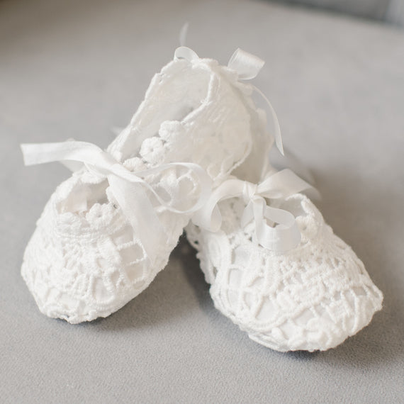 A pair of delicate Grace Lace Booties with silky ribbon ties, resting on a soft grey surface. The intricate lace pattern adds a touch of elegance to these tiny shoes.