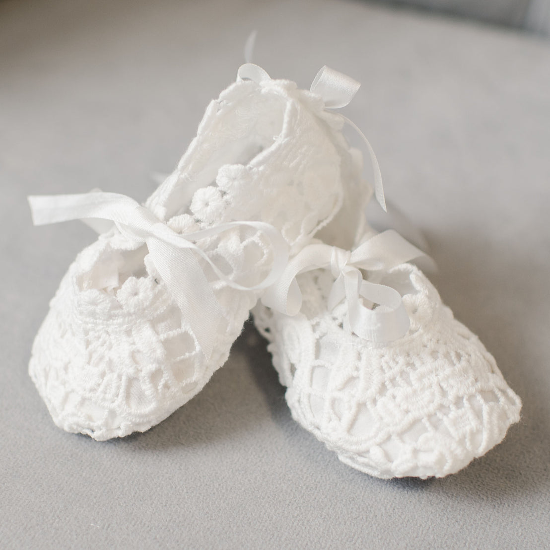 A pair of delicate white lace Grace Shoes with ribbon ties, perfect as a baptism accessory set or baby shower gift, on a soft gray background.