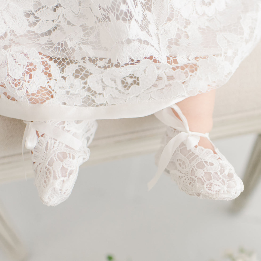 Close-up of a baby's feet wearing Rose Booties, tied with satin ribbons, with the edge of an heirloom lace dress visible. The background is subtly blurred.
