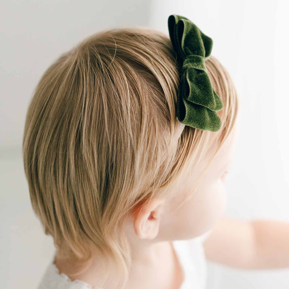 Close-up of a child's head from behind, showcasing short blonde hair neatly styled with an Emily Green Velvet Bow Headband on top for a baptism. The background is a soft white, emphasizing the simplicity and innocence.