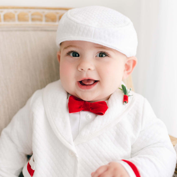 Sentence with product name: A cheerful baby in a white Noah White Newsboy Cap and a red bow tie, ready for a baptism, smiles brightly in a soft and neutral background.
