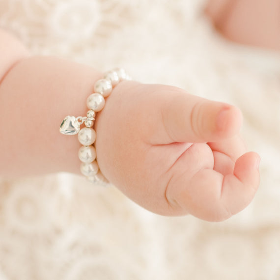 Close-up of a baby's tiny hand with a Baby Beau & Belle White Luster Pearl Bracelet with Silver Heart Charm, resting on a soft, textured blanket.