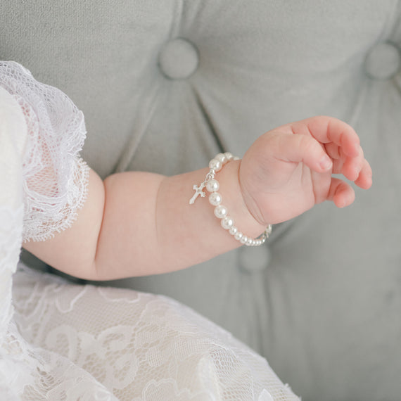 Close-up of a baby's arm wearing a White Luster Pearl Bracelet with Silver Cross. She is wearing the Aria Bubble Romper and sitting on a soft grey sofa.
