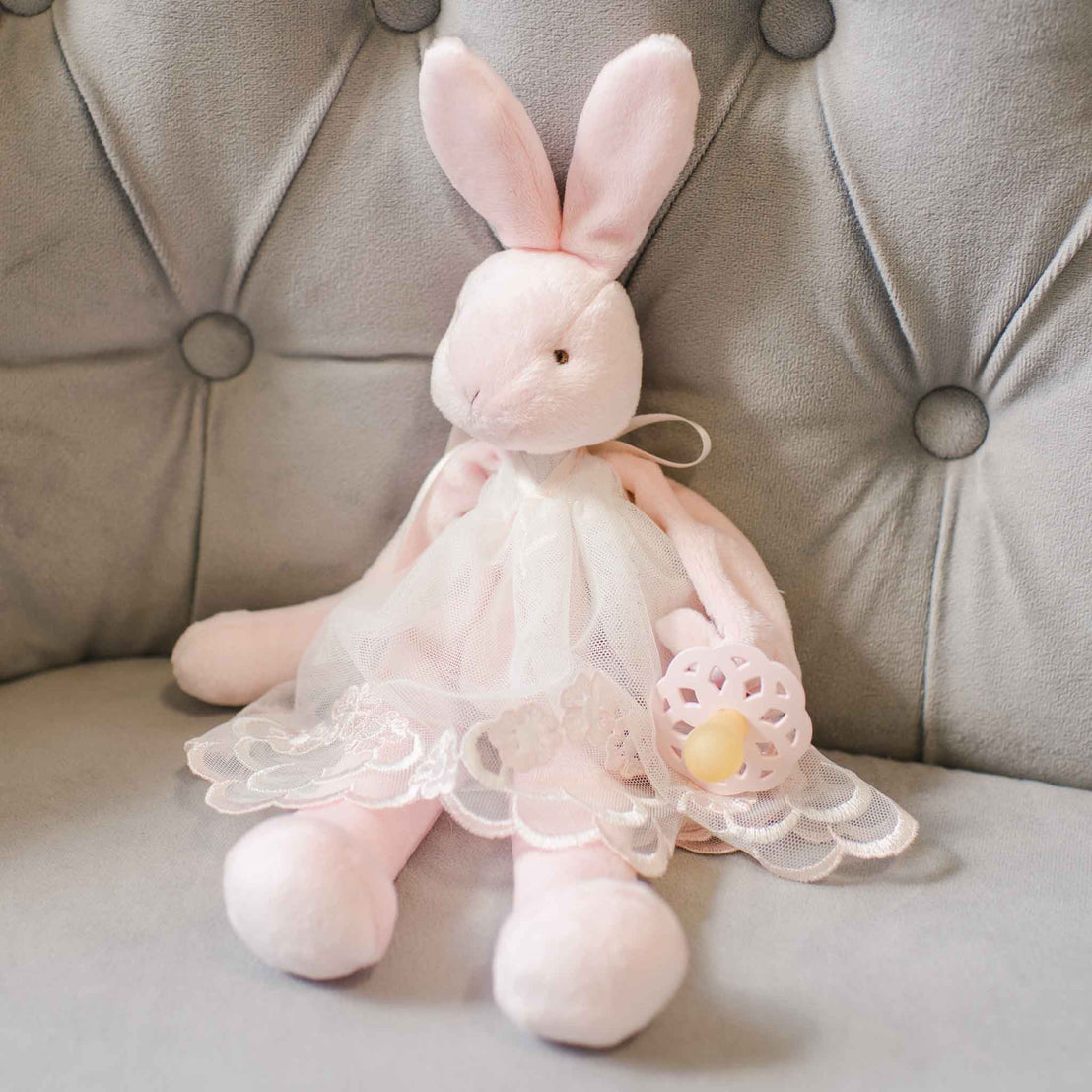 A Joli Silly Bunny Buddy Pacifier Holder dressed in a delicate white lace dress sits on a gray tufted sofa, holding a vintage-inspired yellow and pink Easter egg.