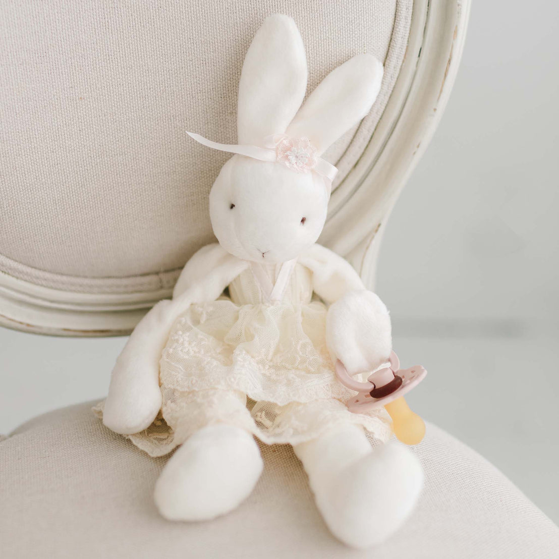 A Jessica Silly Bunny Buddy Pacifier Holder dressed in a lacy dress and holding a Pacifier, sitting elegantly on a beige chair, creating a serene and soft ambiance.