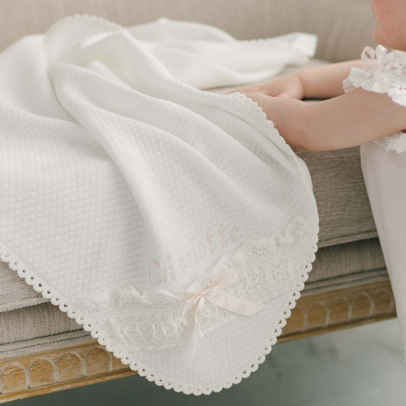 A baby's legs peeking out from under a Charlotte Personalized Blanket, resting on a gray couch. The blanket features lace and ribbon details suitable for.