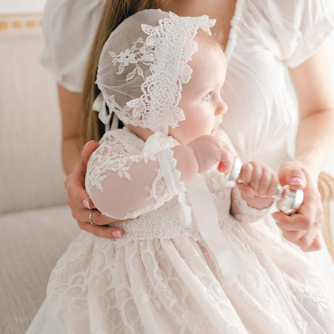 A mother in a white lace dress gently holds her baby, dressed in a Juliette Christening Gown & Bonnet, focusing tenderly on her child.