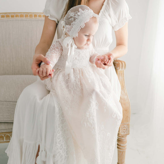 A mother in a white dress holds her baby, dressed in a delicate handcrafted Juliette Christening Gown & Bonnet, as they sit on a beige sofa in a bright room.
