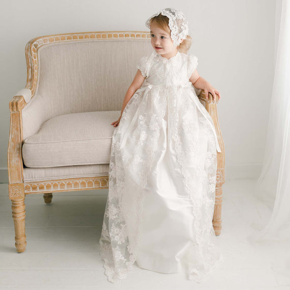 Baby girl sitting in the full-length Penelope christening gown made of super-soft silk and luxurious lace netting.