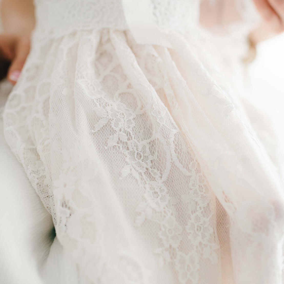 Close-up of a bride's Juliette Christening Gown & Bonnet, focusing on the detailed floral patterns on the vintage fabric, with a soft light illuminating the gown.