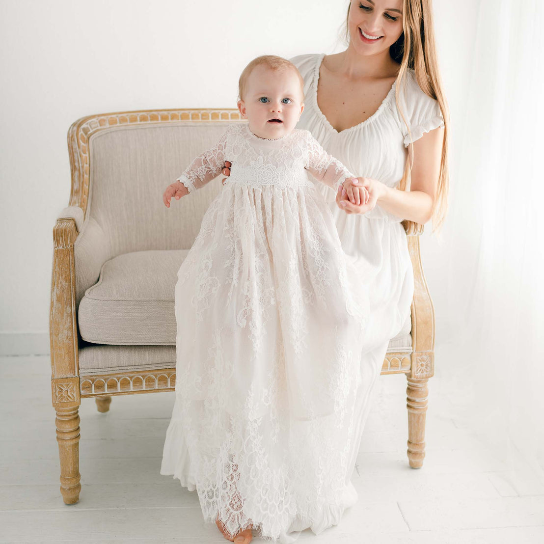 A mother, smiling, helps her baby daughter in a Juliette Christening Gown & Bonnet take her first steps beside an upscale chair in a bright room.