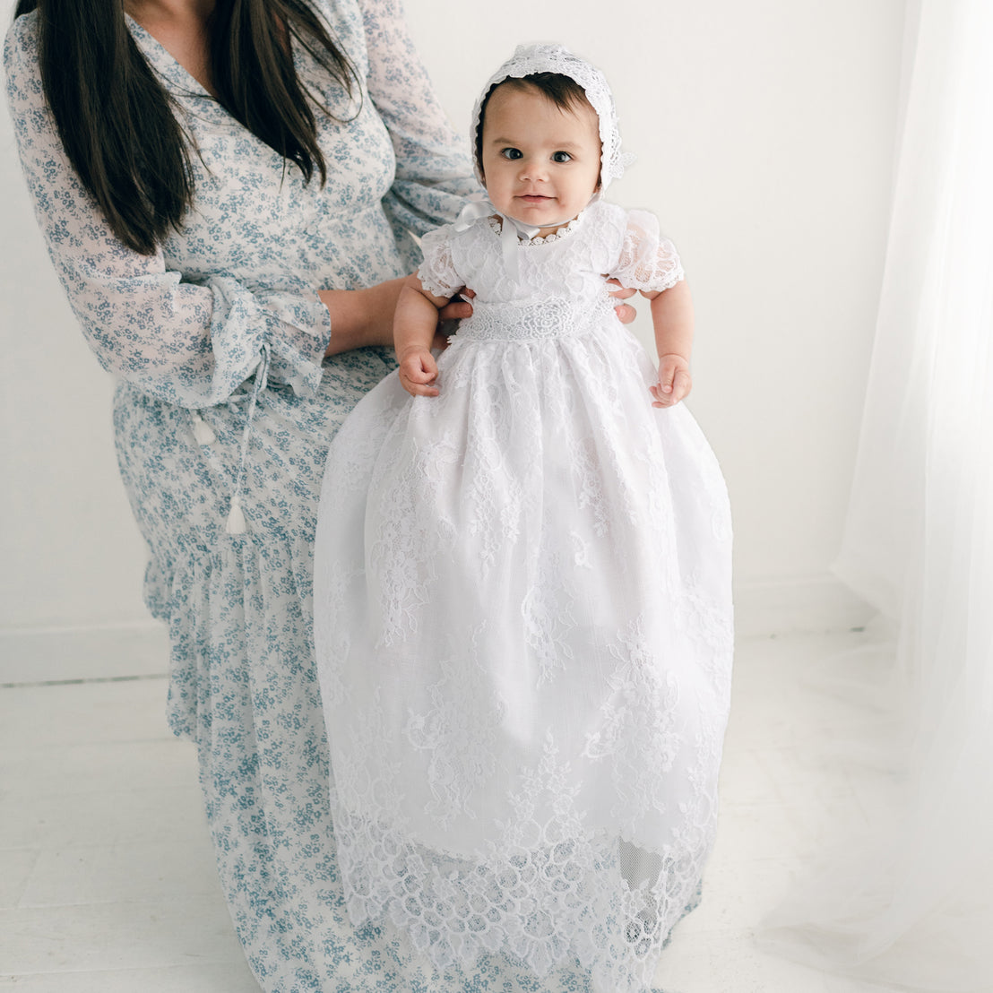 A mother holding her baby, dressed in the Olivia Christening Gown & Bonnet, with the baby looking directly at the camera. They stand against a plain white backdrop.