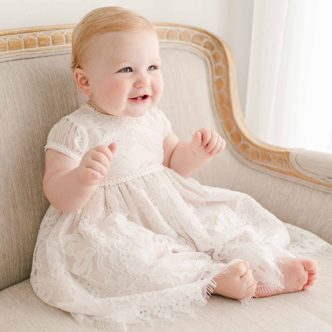 Smiling baby girl sitting and wearing the pink Victoria Puff Sleeve Christening Dress. The dress made with an embroidered ivory lace and lined with pink silk Dupioni.
