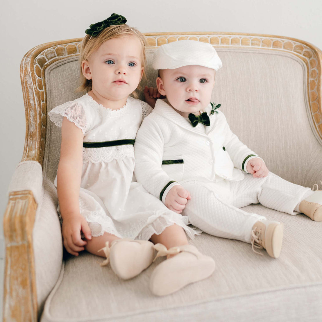 A toddler girl and a baby boy dressed in formal white outfits, sitting together on a beige sofa for their christening. The girl has a green headband, and the boy sports a Noah 3-Piece Suit | Green Trim.