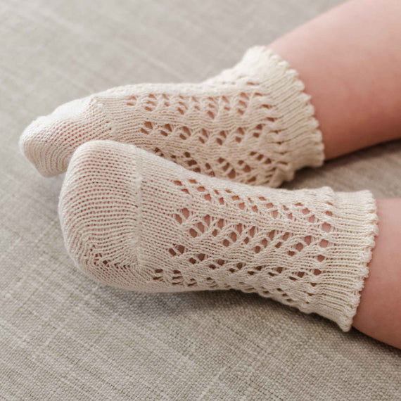 A close-up of a baby’s feet wearing delicate, Crochet Socks in cream lace. The texture of the socks contrasts with the soft, plush surface beneath.