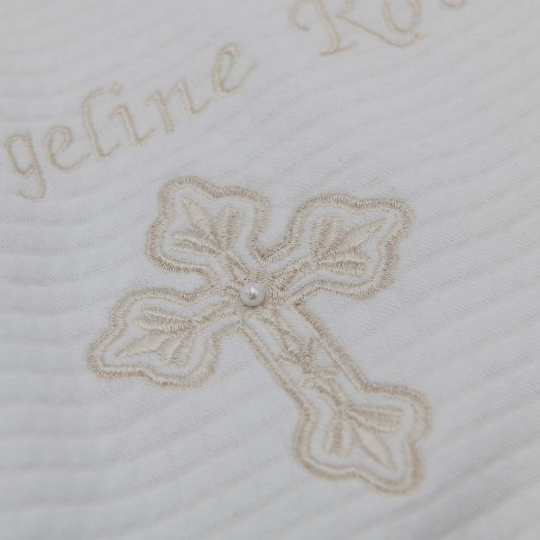 Close-up of a white Personalized Cross Blanket embroidered with the name "angeline rose" in cursive and a detailed cross design with a pearl at the center, used for baptism or christening