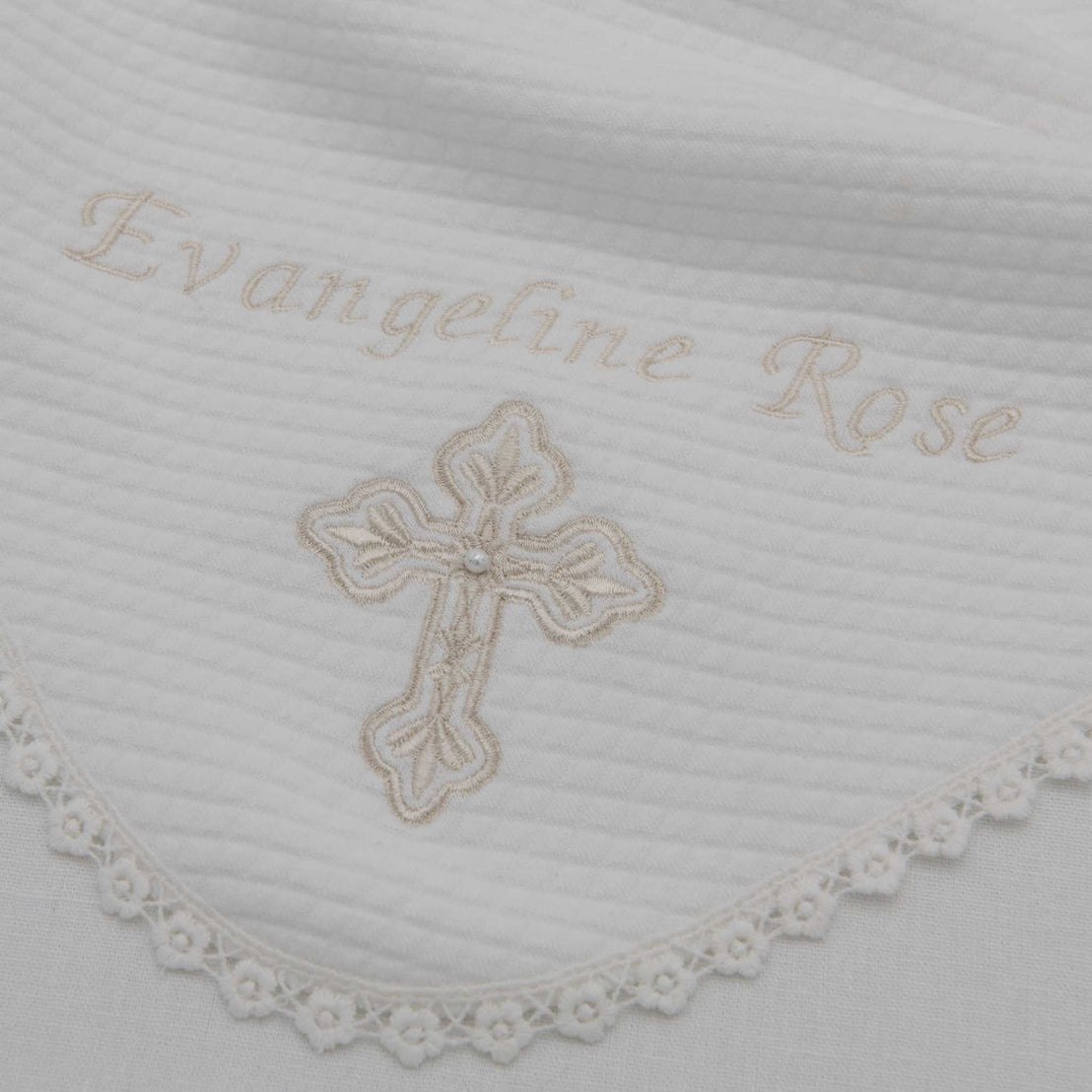 A close-up of a white fabric with the embroidered name "evangeline rose" and a delicate gold cross for a christening, surrounded by a lace trim edge on the Personalized Cross Blanket.