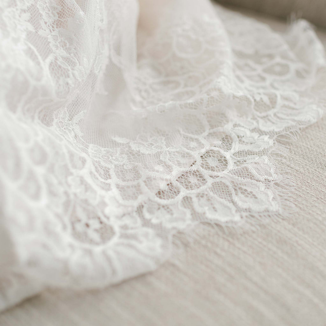 Close-up of delicate Juliette Christening Gown & Bonnet lace fabric with intricate floral patterns, displayed on a soft, vintage, textured surface.