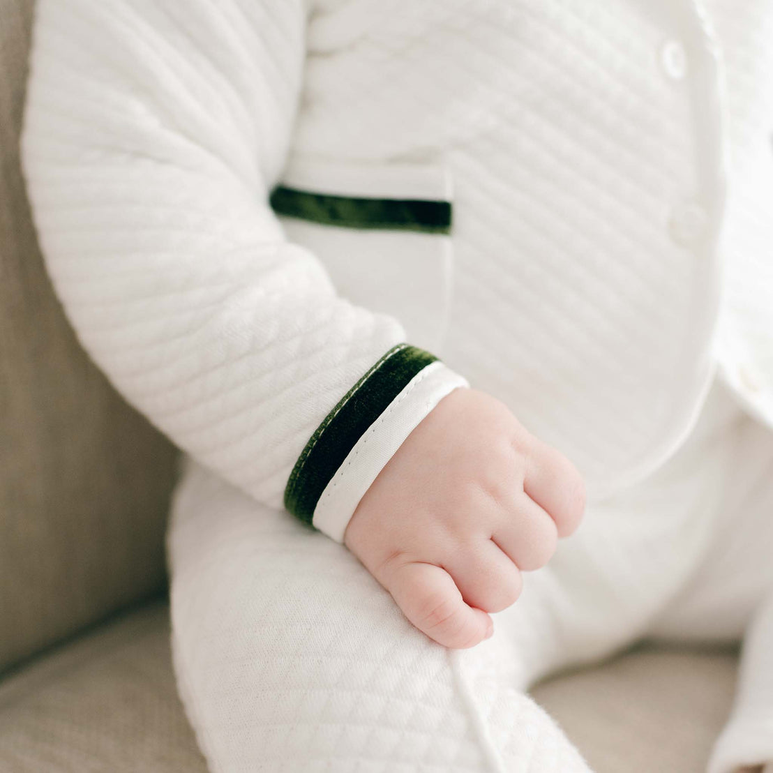 A close-up photo of a baby's fist wearing a Noah 3-Piece Suit with green trim, seated on an upscale, soft light-colored surface.