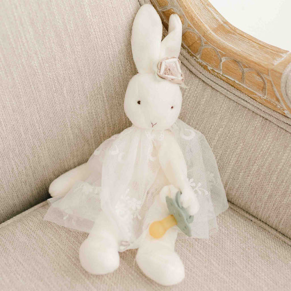 A soft, plush Eliza Silly Bunny Buddy in a vintage tulle dress sitting on a beige upholstered chair, holding a small, blue pacifier. The bunny has a gentle smile and a bow on one ear.