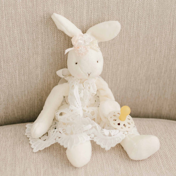 Flat lay of the Lola Silly Bunny Buddy, a floppy stuffed animal bunny that can velcro onto a pacifier. The bunny is dressed to match the Lola gown, dress, and romper with a lace dress and beaded flower headband.