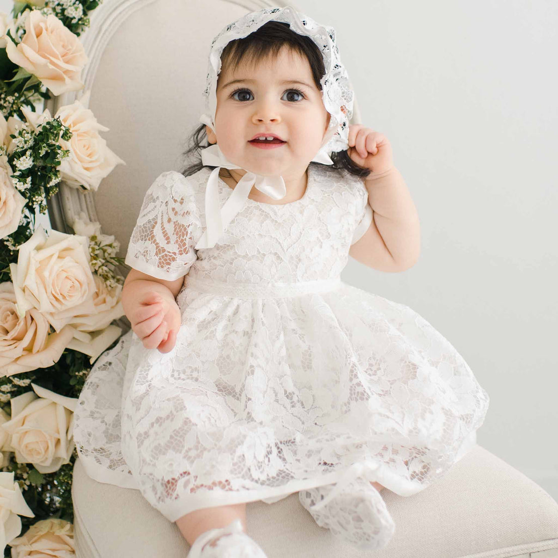 A young child wearing a white lace Rose Romper Dress and bonnet sits on a cream chair, surrounded by pink roses, looking upward with a joyful expression at their baptism.