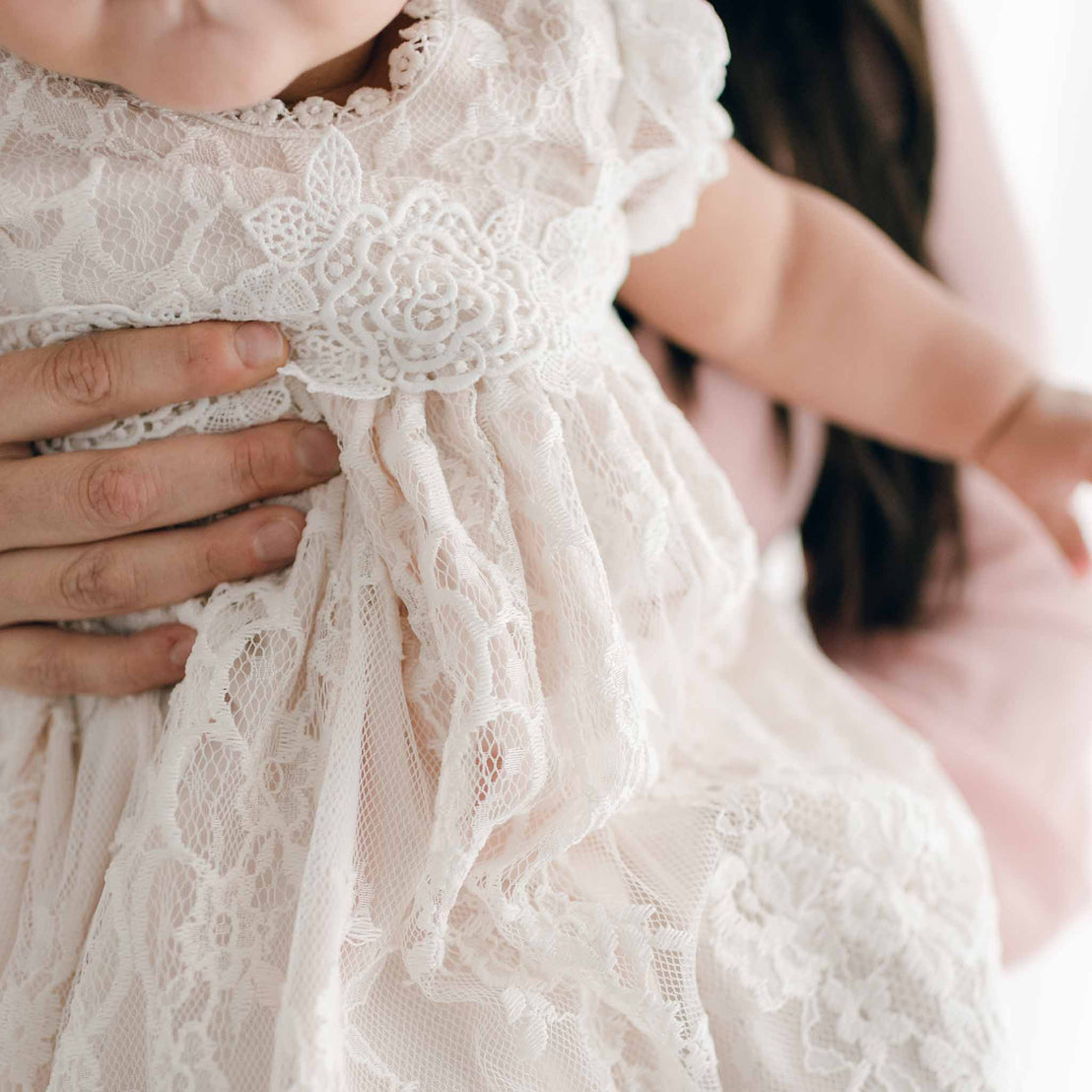 Close-up of a woman holding a child dressed in a Juliette Layette, focusing on the intricate fabric and tender embrace against a soft, light background.