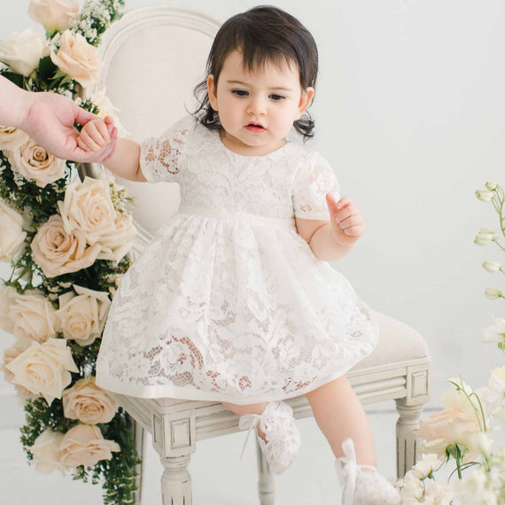 A toddler in a Rose Romper Dress sits on a traditional vintage chair, holding an adult's hand, surrounded by a soft floral backdrop.
