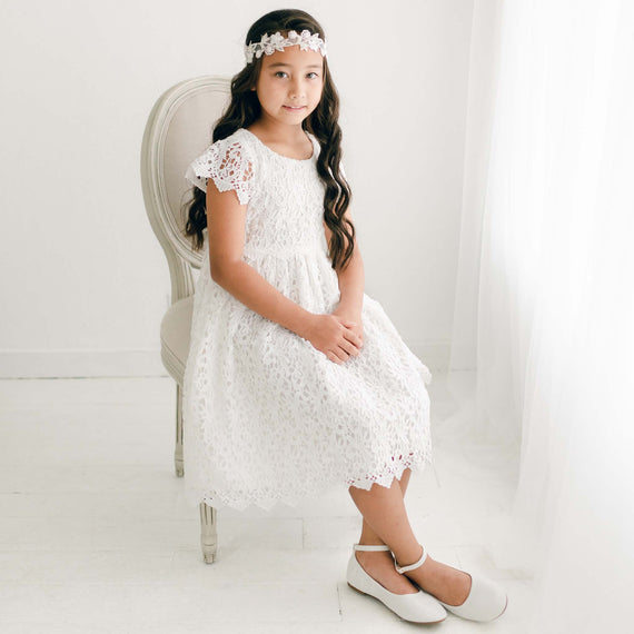 An adolescent girl wearing the Lola Girls Lace Dress. Made with cotton lining in light ivory and adorned with an all-over embroidered lace, plus a lace bodice, lacey cap sleeves, and buttons in back. She is also wearing the Lola velvet headband.
