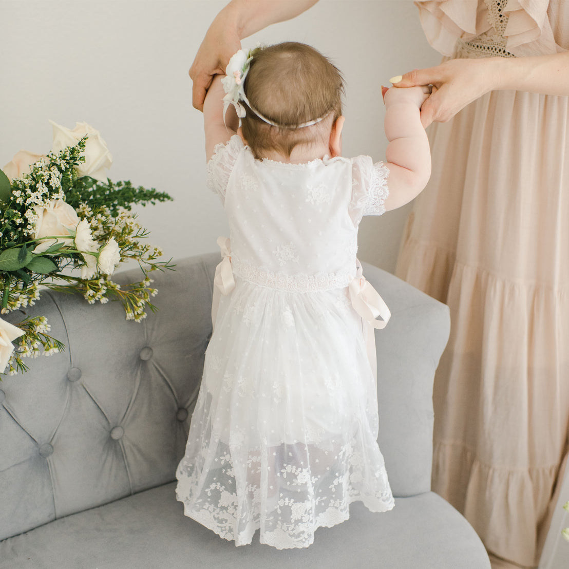 A baby in a Melissa Romper Dress is held up by her mother, who is adjusting a floral headband on her head, in a softly decorated room with flowers nearby.
