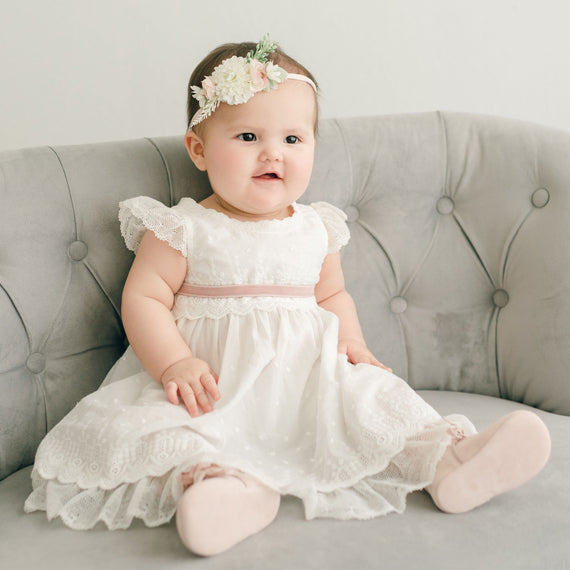 Baby girl wearing the mauve Emily Dress, Bloomers, Sash Tie and Emily Flower Headband. The dress features embroidered cotton eyelet lace with a scalloped edge, lace trim and soft lace cap sleeves.