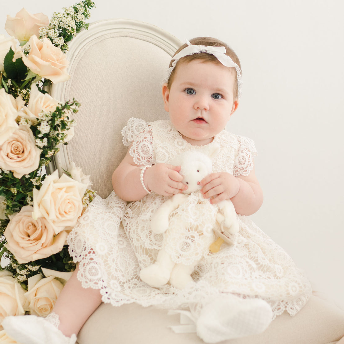 Baby girl sitting in chair holding a soft toy bear. Wearing the Poppy fancy baby dress.