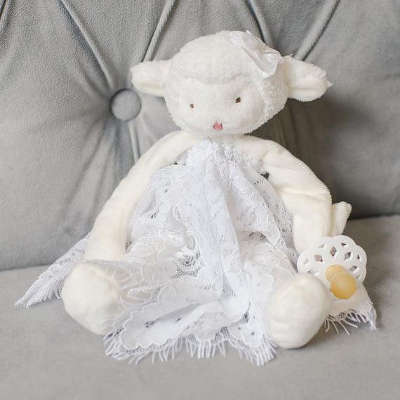 An Aria Silly Lamb Buddy with a lace dress sits on a gray couch holding the Aria Pacifier.