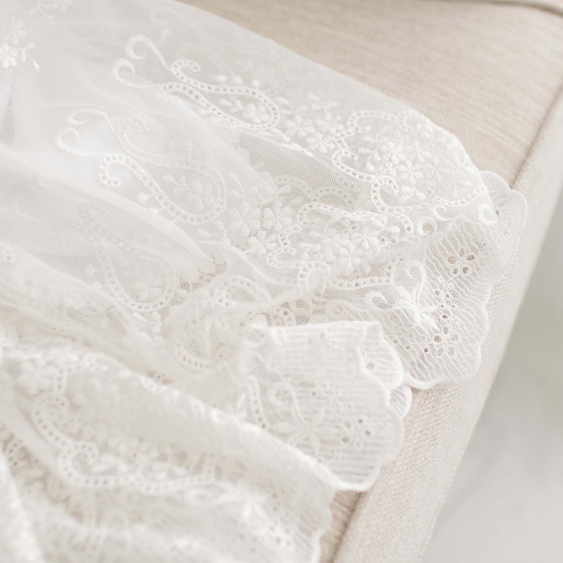 A close-up of the Eliza Blessing Gown draped over a soft beige chair, showcasing intricate floral patterns and embroidery details suitable for a christening or baptism.