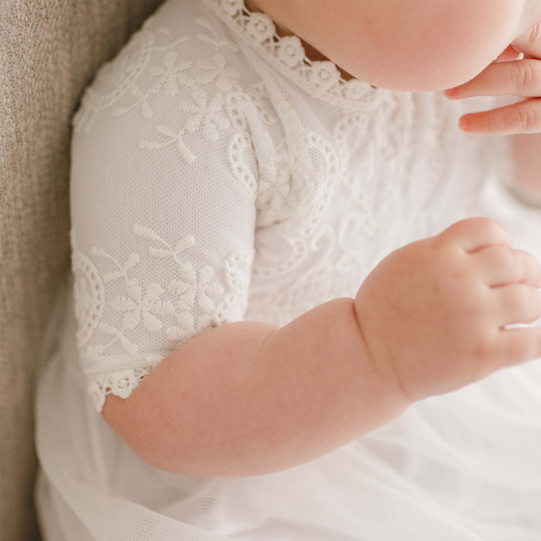 Close-up of a baby dressed in the Eliza Blessing Gown & Bonnet for a baptism, focusing on the delicate fabric details and the chubby arm of the infant against a soft background.