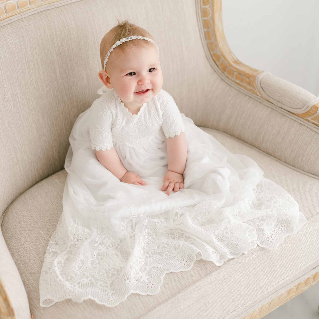 A joyful baby in an Eliza Blessing Gown & Bonnet, prepared for a christening, sits on an upscale beige vintage sofa, smiling broadly. The baby wears a delicate headband, enhancing a sweet, cheerful expression.