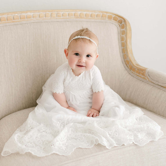 A baby wearing the Eliza Blessing Gown & Bonnet suited for a christening sits on an upscale beige vintage sofa, smiling at the camera.