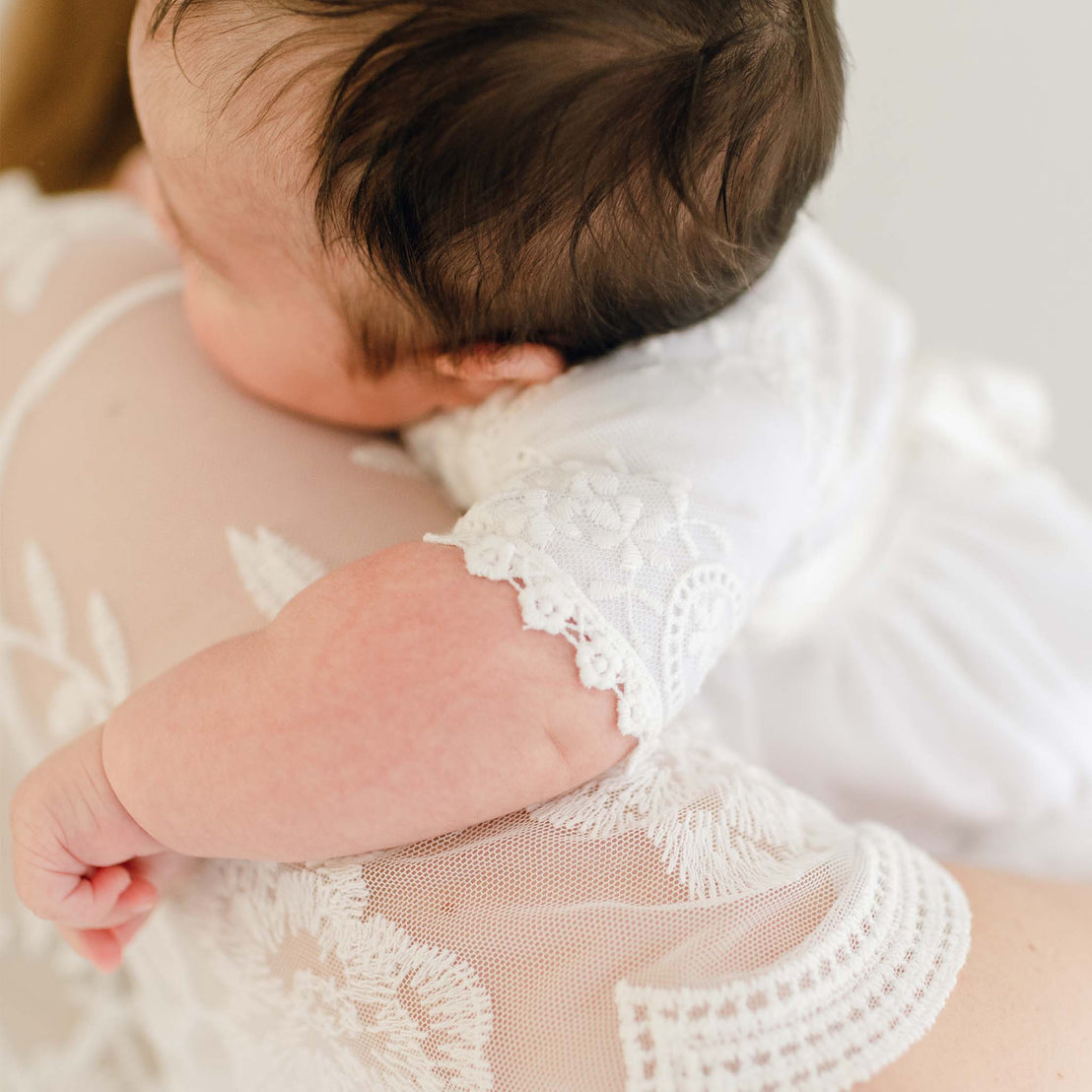 A close-up image of a baby with dark hair being gently held against a mother's shoulder, who is wearing the Eliza Blessing Gown & Bonnet. The focus is on the baby’s tiny hand and delicate textures.