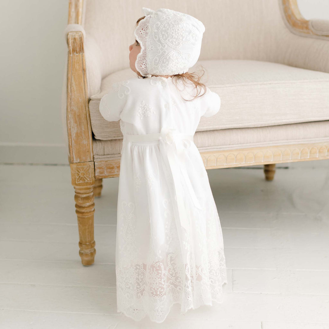 A toddler in the Eliza Lace Dress & Headband stands by an elegant antique chair, facing away from the camera. The setting is a bright room with soft-colored decor.